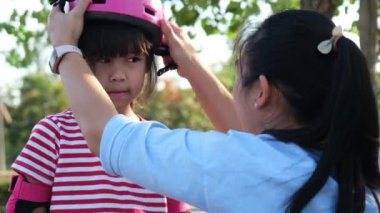 Young mother helps her daughter put on her protection pads and helmet before roller skating in the park. Active leisure and outdoor sport for child.