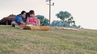 Happy sisters playing at the park slides down from the grassy hill sitting on a cardboard box. Happy children playing outdoors in summer. Family spending time together on vacation.