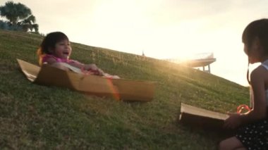 Happy sisters playing at the park slides down from the grassy hill sitting on a cardboard box. Happy children playing outdoors in summer. Family spending time together on vacation.