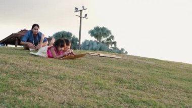 Happy sisters playing at the park slides down from the grassy hill sitting on a cardboard box. Happy children playing outdoors in summer. Family spending time together on vacation.