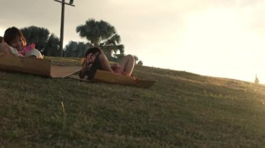 Happy sisters playing at the park slides down from the grassy hill sitting on a cardboard box. Happy children playing outdoors in summer. Family spending time together on vacation.