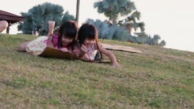 Happy sisters playing at the park slides down from the grassy hill sitting on a cardboard box. Happy children playing outdoors in summer. Family spending time together on vacation.