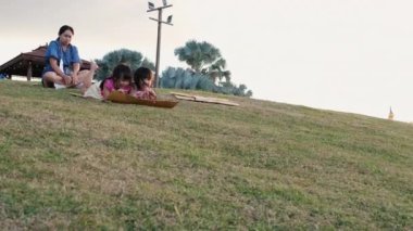 Happy sisters playing at the park slides down from the grassy hill sitting on a cardboard box. Happy children playing outdoors in summer. Family spending time together on vacation.