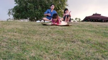 Happy sisters playing at the park slides down from the grassy hill sitting on a cardboard box. Happy children playing outdoors in summer. Family spending time together on vacation.