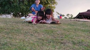 Happy sisters playing at the park slides down from the grassy hill sitting on a cardboard box. Happy children playing outdoors in summer. Family spending time together on vacation.