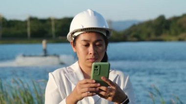 Environmental engineer wearing a white helmet uses a mobile phone to record data analyzing oxygen levels in a reservoir. Water and ecology concept.