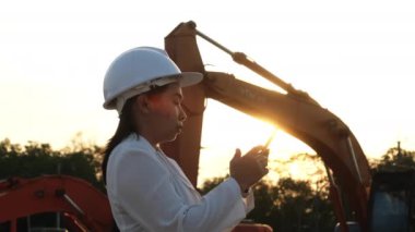 Portrait of an Asian engineer wearing a white helmet using a smartphone in a workplace. Civil engineer checking after work on construction site during sunset on yellow excavator background.