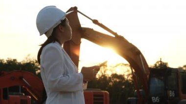 Portrait of an Asian engineer wearing a white helmet using a smartphone in a workplace. Civil engineer checking after work on construction site during sunset on yellow excavator background.
