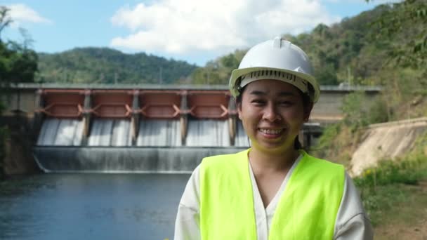 水力発電ダムを背景に外に立つ緑のベストとヘルメットで笑顔の女性エンジニア 門を通って流れる水と水門 — ストック動画