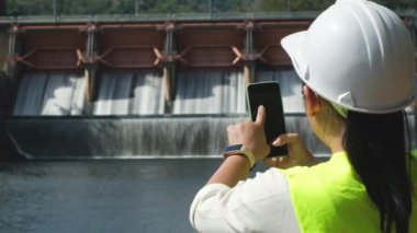 Female engineer in white hat working with smartphone and looking away at dam with hydroelectric power plant and irrigation.