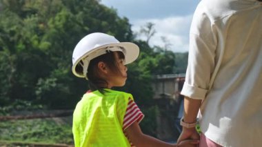 Engineer mother holding her daughter's hand and smiling at each other against the background of a dam with a hydroelectric power station. Concepts of renewable energy and love of nature and family.