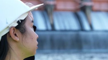 Close-up portrait of a female engineer in a green vest and helmet standing outside against the background of a dam with a hydroelectric power station and irrigation.