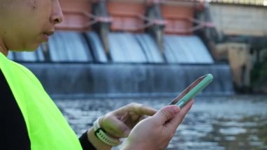Female environmentalist wearing green vest and safety helmet working outdoors against the background of hydroelectric dam and recording analytical data for water contamination on mobile phone.