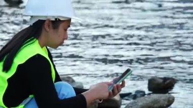 Female environmentalist using mobile phone to record water analysis data in dam. Researchers are collecting water samples from the river to test and detect pathogens. Water and ecology concept