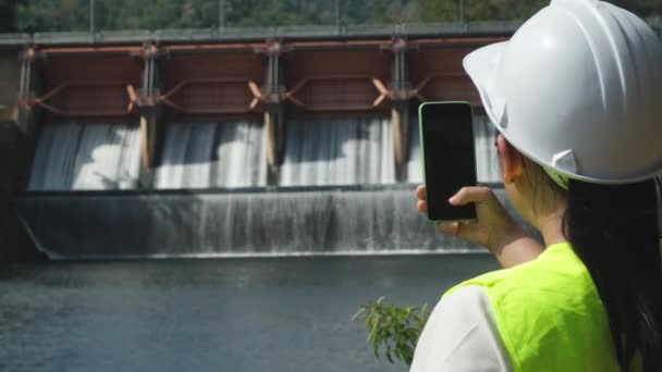 スマートフォンで作業し 水力発電所や灌漑とダムを離れて見て白い帽子の女性エンジニア — ストック動画