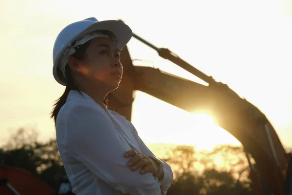 Portrait of Asian engineer wearing white helmet working and inspecting in workplace. Civil engineer checking after work on construction site during sunset on yellow excavator background.