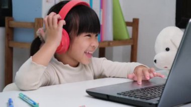 Kulaklık takan ve dizüstü bilgisayar kullanan tatlı bir ilkokul öğrencisi. Mutlu Asyalı çocuklar online olarak bilgisayar ya da evde eğitim, müzik dinlemek ya da oyun oynamak ile çalışıyorlar..