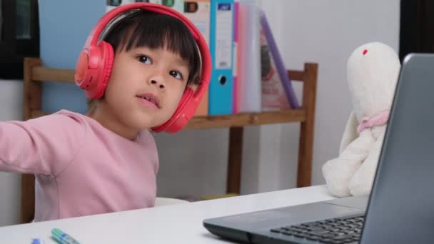 ヘッドフォンをしてノートパソコンを使ってかわいい小学生の女の子 幸せなアジアの子供たちは 音楽を聴いたりゲームをしたりしながら ラップトップコンピュータやホームスクーリングとオンラインで対話的に勉強します — ストック動画