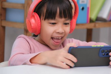 Kulaklık takıp akıllı telefon tutan tatlı bir ilkokul öğrencisi. Akıllı telefondan ya da evde eğitim gören, müzik dinleyen ya da oyun oynayan mutlu Asyalı çocuk..