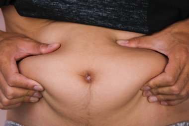 Şişman erkek el, pembe arka planda izole edilmiş göbek yağını tutuyor. Adam şişko karnına dokunuyor. Koca göbekli şişman genç bir adam.