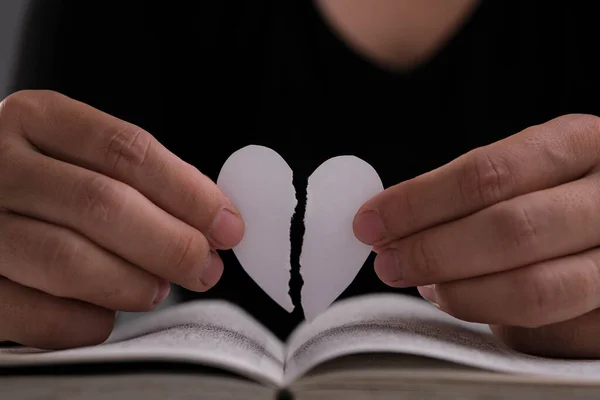 テーブルの上に開かれた本の上に半分裂けた白い紙の心を持つ女性の手 白い紙の心の手で半分に引き裂かれた — ストック写真