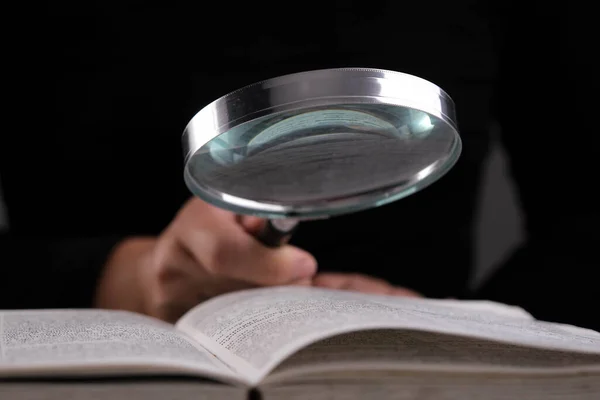 Close-up of a woman looking through a magnifying glass at a textbook. Magnifying glass in hand and open book on table. Education and research concept.
