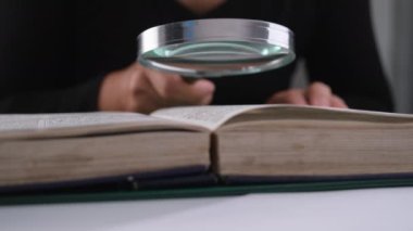 Ders kitabına büyüteçle bakan bir kadının yakın çekimi. Elde büyüteç ve masada açık bir kitap. Eğitim ve araştırma kavramı.