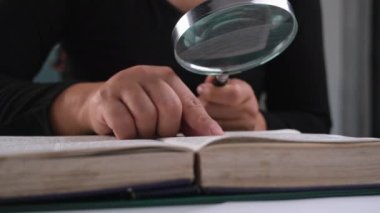 Ders kitabına büyüteçle bakan bir kadının yakın çekimi. Elde büyüteç ve masada açık bir kitap. Eğitim ve araştırma kavramı.