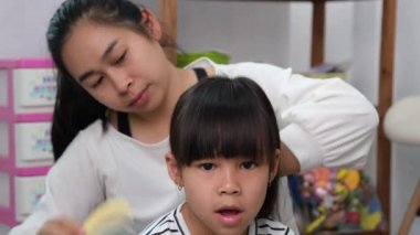 Güzel annesi kızının saçını tararken sevimli küçük kız gülümsüyor. Anne odada oturmak için kızının saçını tarıyor. Mutlu aile ve Anneler Günü konsepti.