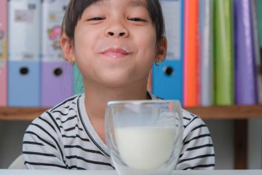 Oturma odasında bir bardak süt içen tatlı Asyalı kız. Sabah okula gitmeden önce süt içen küçük bir kız. Çocuklukta sağlıklı yiyecekler.