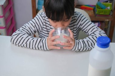 Oturma odasında bir bardak süt içen tatlı Asyalı kız. Sabah okula gitmeden önce süt içen küçük bir kız. Çocuklukta sağlıklı yiyecekler.