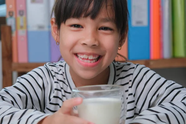 stock image Cute Asian girl drinking a glass of milk at home in living room. Little girl drinking milk in the morning before going to school. Healthy food in childhood.