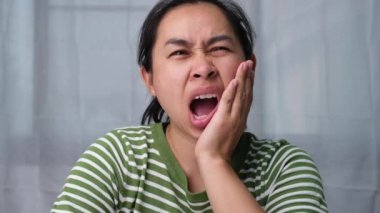 Elini yanağında tutan Asyalı kadın diş ağrısı, diş çürüğü, diş eti iltihabı ya da kötü nefes problemi hissediyor. Diş ağrısı ve oral sağlık konsepti.