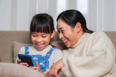 Genç anne ve küçük kızı akıllı telefon kullanıyor ve evdeki kanepede otururken gülümsüyorlar. Annesiyle evde dinlenirken akıllı telefon kullanan neşeli küçük kız. Çocuklar ve Teknoloji