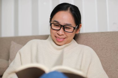 Kanepede oturup içeride kitap okuyan mutlu Asyalı kadın. Beyaz kazaklı bir kadının portresi evdeki kanepede kitap okuyor. Yaşam tarzı, boş zaman ve rahatlama.