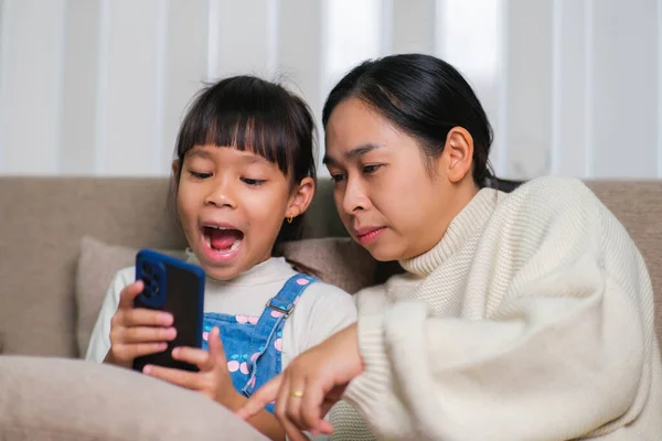 年轻的母亲和她的小女儿在家里坐在沙发上 一边用智能手机一边笑 快乐的小女孩在家里和妈妈放松的时候用智能手机 儿童与技术 — 图库照片