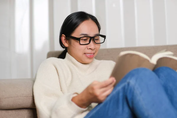 幸せなアジアの女性はソファに座って本を屋内で読んでいます 白いセーターの女性の自宅でソファの上で本を読んで肖像画 ライフスタイル 自由な時間とリラクゼーション — ストック写真