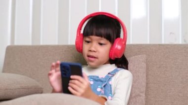 Sıradan kıyafetler ve kulaklıklar takan akıllı telefon kullanan ve odada müzik dinleyen tatlı küçük bir kız. Kablosuz kulaklıklı ve cep telefonlu gülümseyen kız..