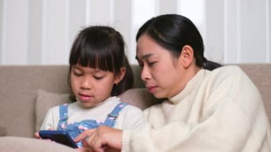 Genç anne ve küçük kızı akıllı telefon kullanıyor ve evdeki kanepede otururken gülümsüyorlar. Annesiyle evde dinlenirken akıllı telefon kullanan neşeli küçük kız. Çocuklar ve Teknoloji