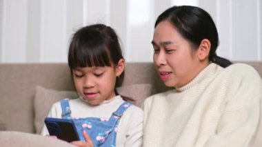 Genç anne ve küçük kızı akıllı telefon kullanıyor ve evdeki kanepede otururken gülümsüyorlar. Annesiyle evde dinlenirken akıllı telefon kullanan neşeli küçük kız. Çocuklar ve Teknoloji