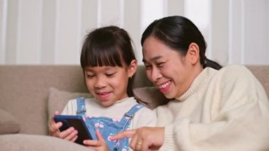 Genç anne ve küçük kızı akıllı telefon kullanıyor ve evdeki kanepede otururken gülümsüyorlar. Annesiyle evde dinlenirken akıllı telefon kullanan neşeli küçük kız. Çocuklar ve Teknoloji