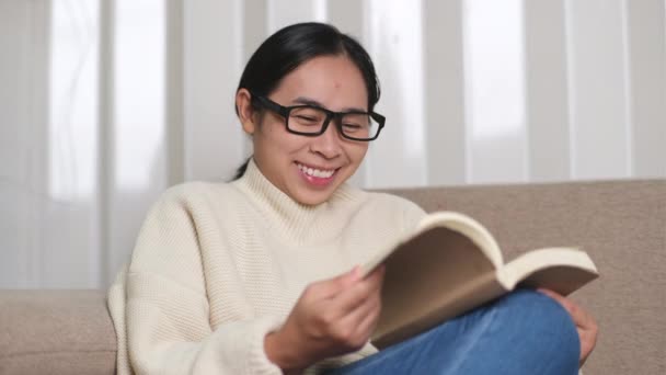 幸せなアジアの女性はソファに座って本を屋内で読んでいます 白いセーターの女性の自宅でソファの上で本を読んで肖像画 ライフスタイル 自由な時間とリラクゼーション — ストック動画