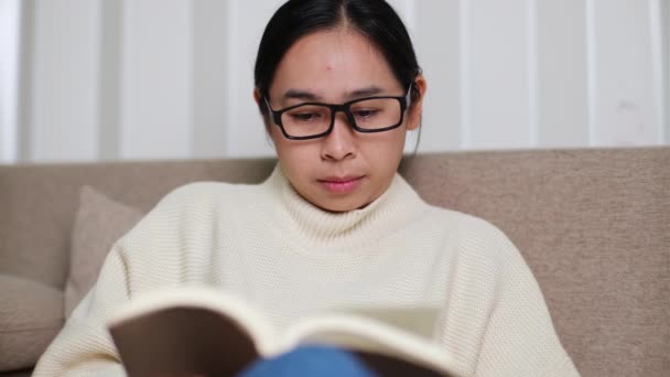 快乐的亚洲女人坐在沙发上 在屋里看书 一个穿着白色毛衣的女人在家里沙发上看书的画像 生活方式 闲暇时间和放松 — 图库视频影像