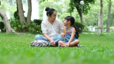 Anne ve küçük kızı parkta çimlerin üzerinde birlikte oturuyorlar. Anne yeşil doğa parkında küçük kızıyla eğleniyor. Mutlu aile kavramı. Anneler Günü
