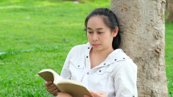 夏の公園で本を読んでジャケットでリラックスした若い女性 女性は草の上に座り 休暇中に木の下で休んでいます 教育理念 — ストック動画
