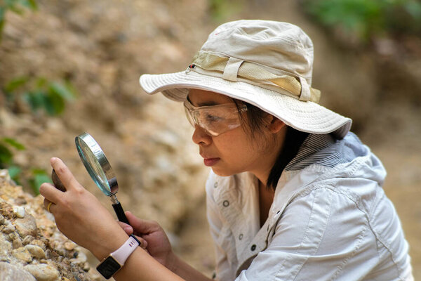 Female geologist using magnifying glass to examine and analyze rock, soil, sand in nature. Archaeologists explore the field. Environmental and ecology research.