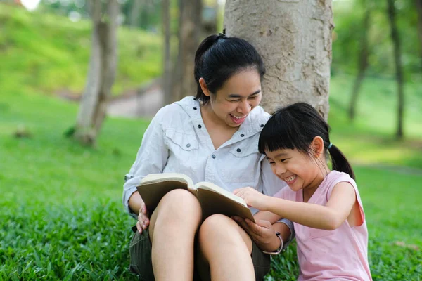 Happy mother and little girl having fun and enjoying reading at the park. Mother and daughter resting in the summer garden, she reads fairy tales to her daughter.