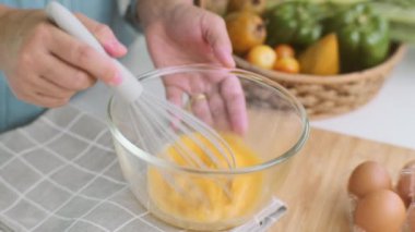 Parlak mutfakta yemek pişiren genç bir kadın, elleri havluya ve ahşap masaya yerleştirilmiş bir kasede yumurta çırpıyor. Sağlıklı yemek için malzemeleri hazırlıyorum. Ev yapımı yemek.