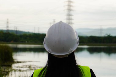 Kadın çevre mühendisi, arka planda elektrik santrali ve yüksek voltaj direklerine karşı bir su deposunun yanında duruyor. Enerji, ekoloji ve çevre kavramı