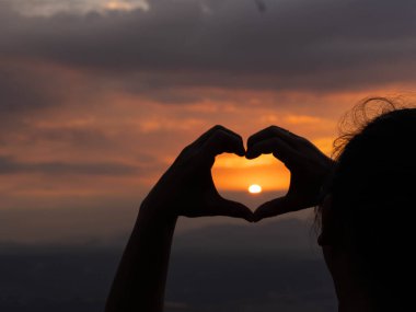 Kadın ellerinin silueti günbatımı arka planında kalp şekli oluşturuyor. Doğa için kalp şeklinde..